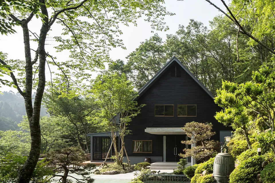 亀岡のVILLA／保養所。木々に囲まれた庭の奥に建つ、大きな切妻屋根と黒い外壁の建物