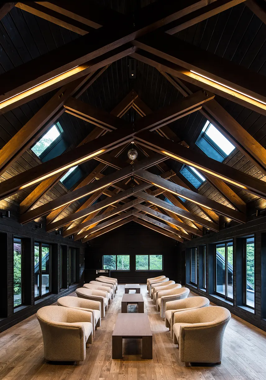 亀岡のVILLA／保養所。照明で照らされた木梁が交差する勾配天井の下に、アームチェアを向かい合わせに並べたラウンジ