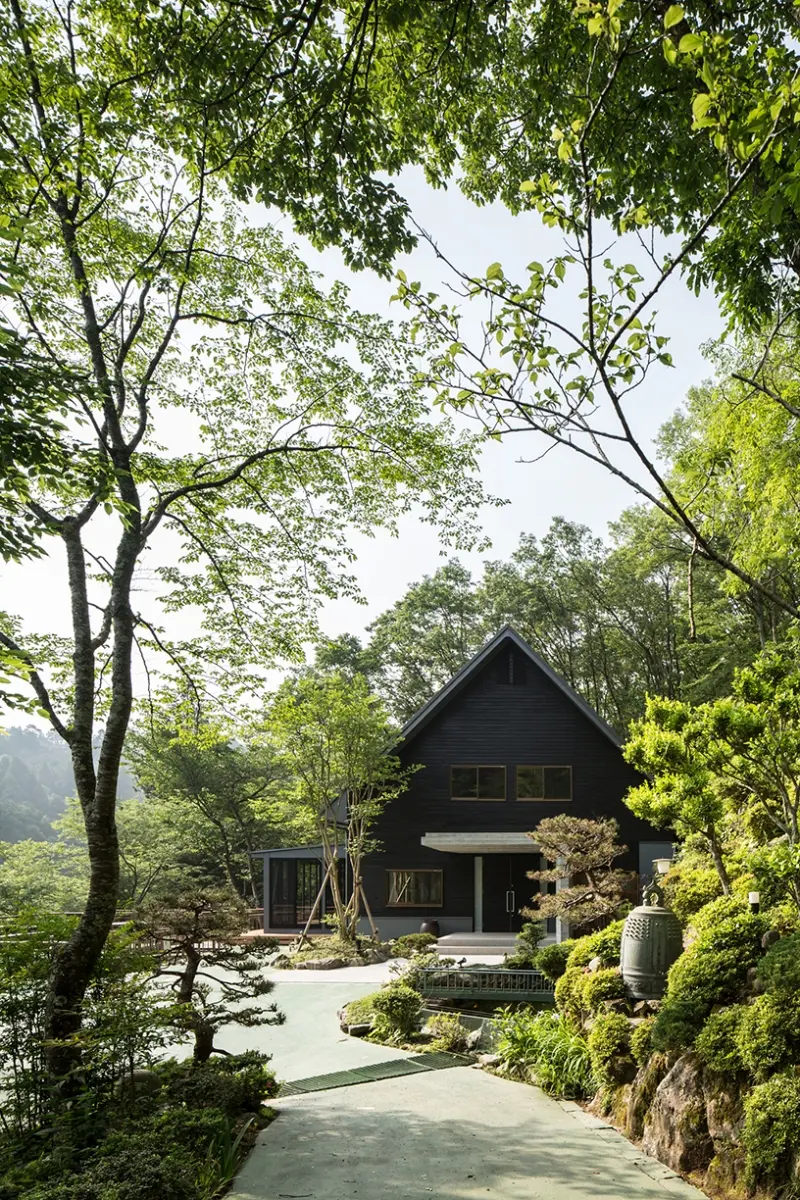 亀岡のVILLA／保養所。木々に囲まれたアプローチの先に、黒い外壁と三角屋根の建物を望む
