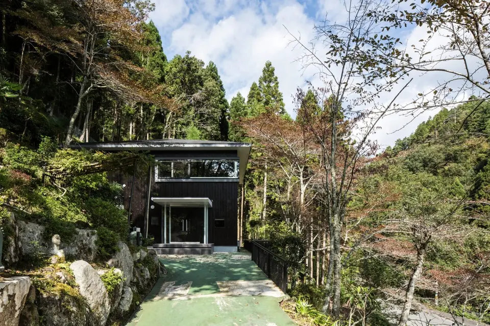 亀岡のVILLA／保養所リノベーション2。木々に囲まれた黒い外壁の建物と、石垣に沿って玄関へ続くアプローチ