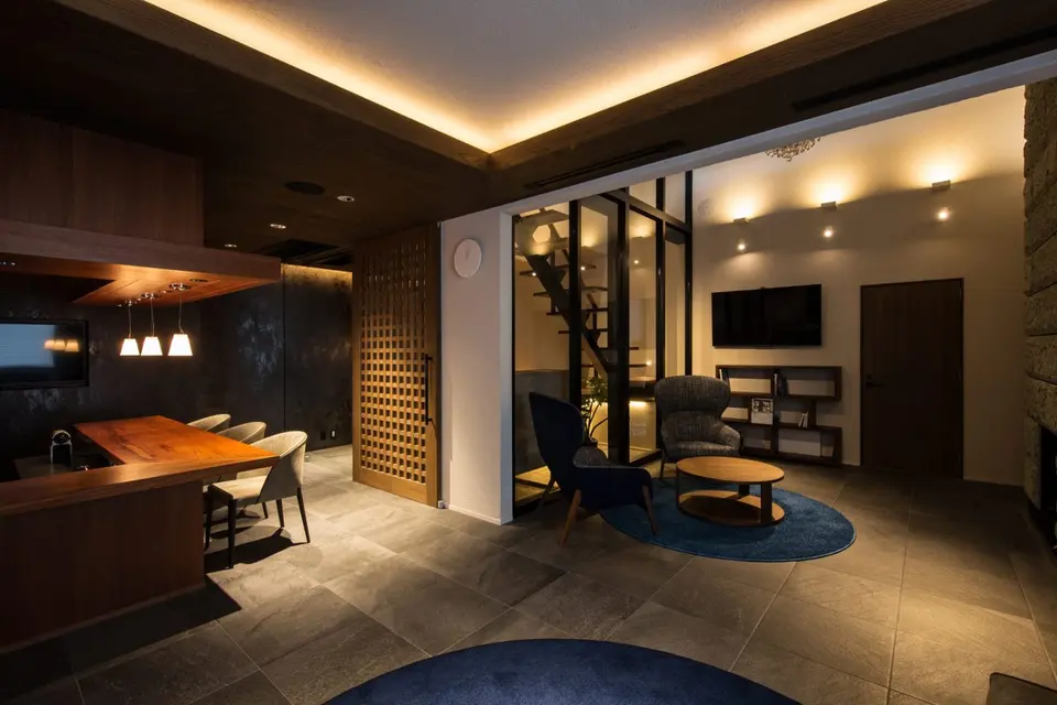 亀岡のVILLA／保養所。間接照明が灯るラウンジ。木のバーカウンターと、ガラス越しに階段が見えるくつろぎのスペース