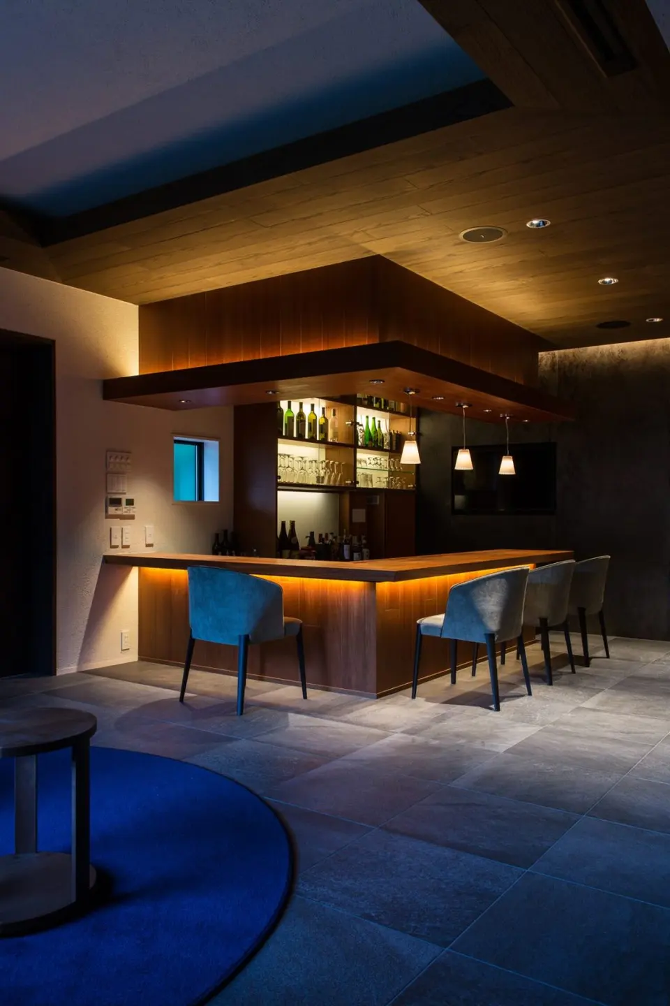 亀岡のVILLA／保養所。木の天井の下、ペンダントライトとカウンター下の照明が灯るL字型のバーカウンター