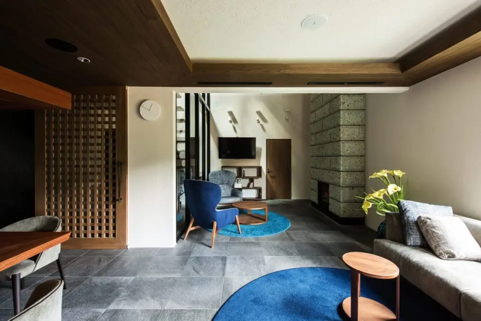 亀岡のVILLA／保養所。グレーのタイル床に青いラグを配し、石張りの暖炉とソファを備えたラウンジ