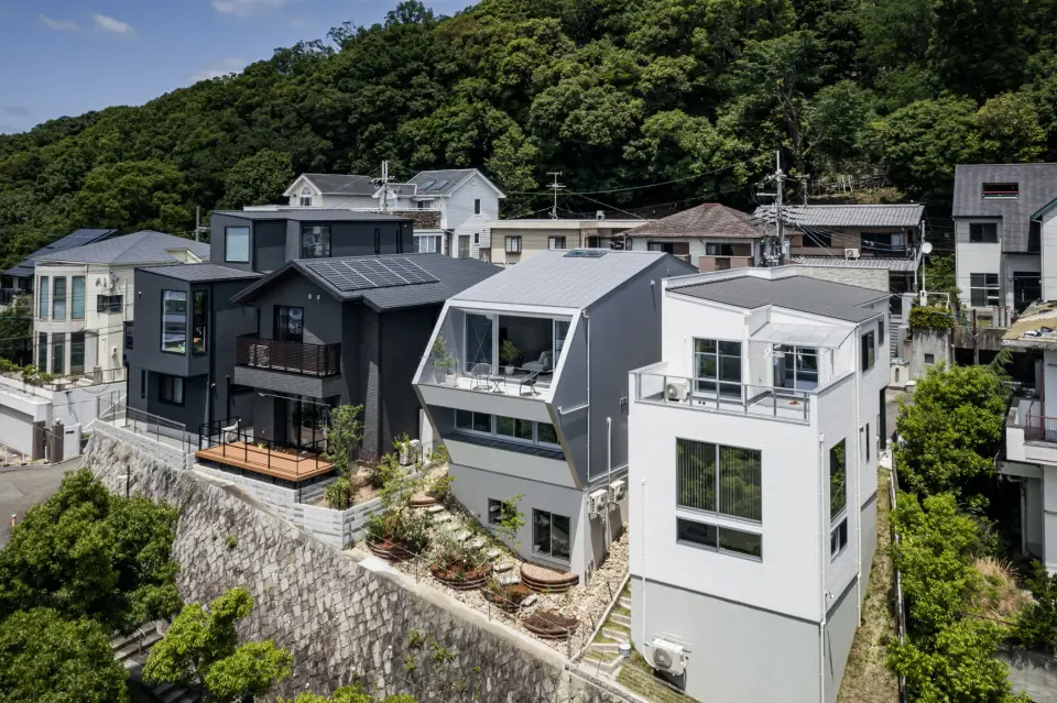五月丘の家。緑の山を背に、石垣の上に並ぶ住宅と、テラスを備えた建物の外観。