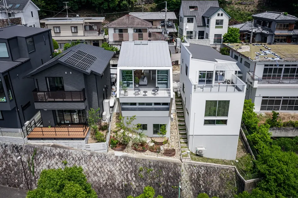 五月丘の家。斜面側の上空から見た住宅。大きな窓とテラス、建物脇をつなぐ外階段。