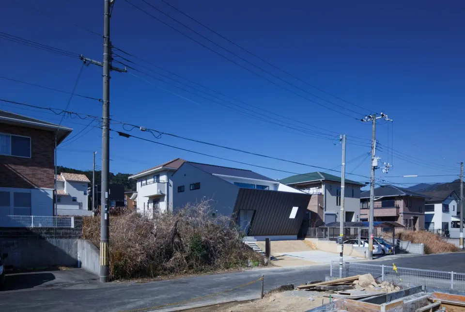 ／（スラッシュ）。住宅街の道路越しに望む、斜めの外壁と白い上階を組み合わせた外観