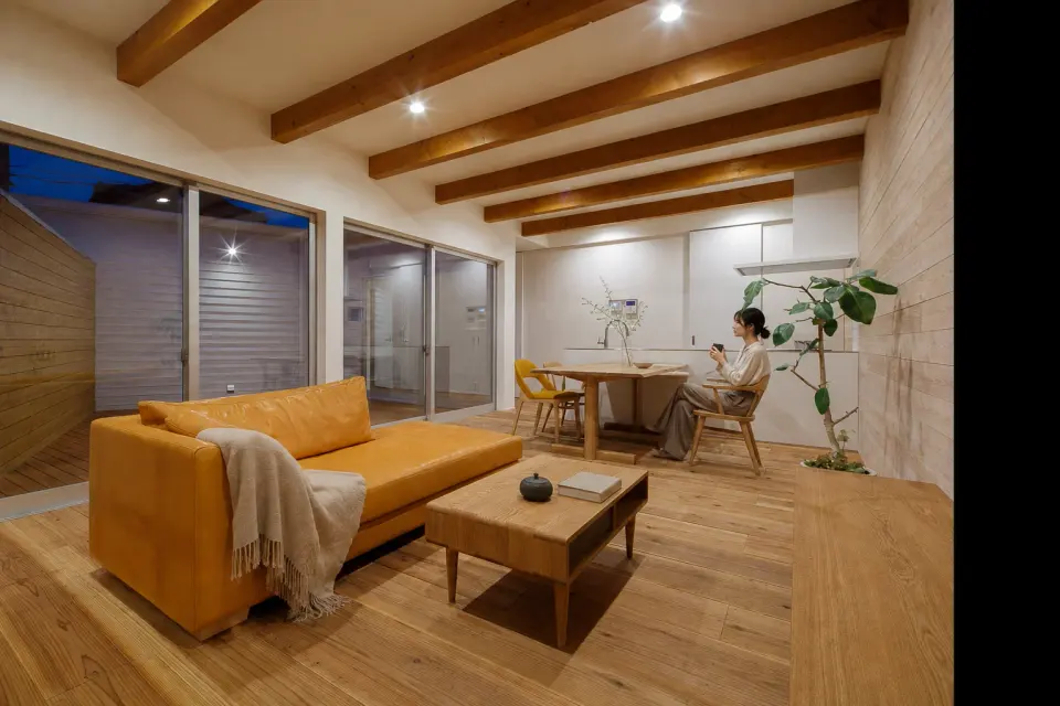 白のコートハウス。木梁と板張りの壁、黄色いソファを配し、奥のダイニングキッチンへ続くリビング