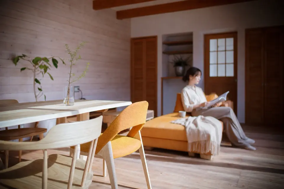 白のコートハウス。木の壁と家具に囲まれたリビングダイニングで、黄色いソファに座って読書をする様子